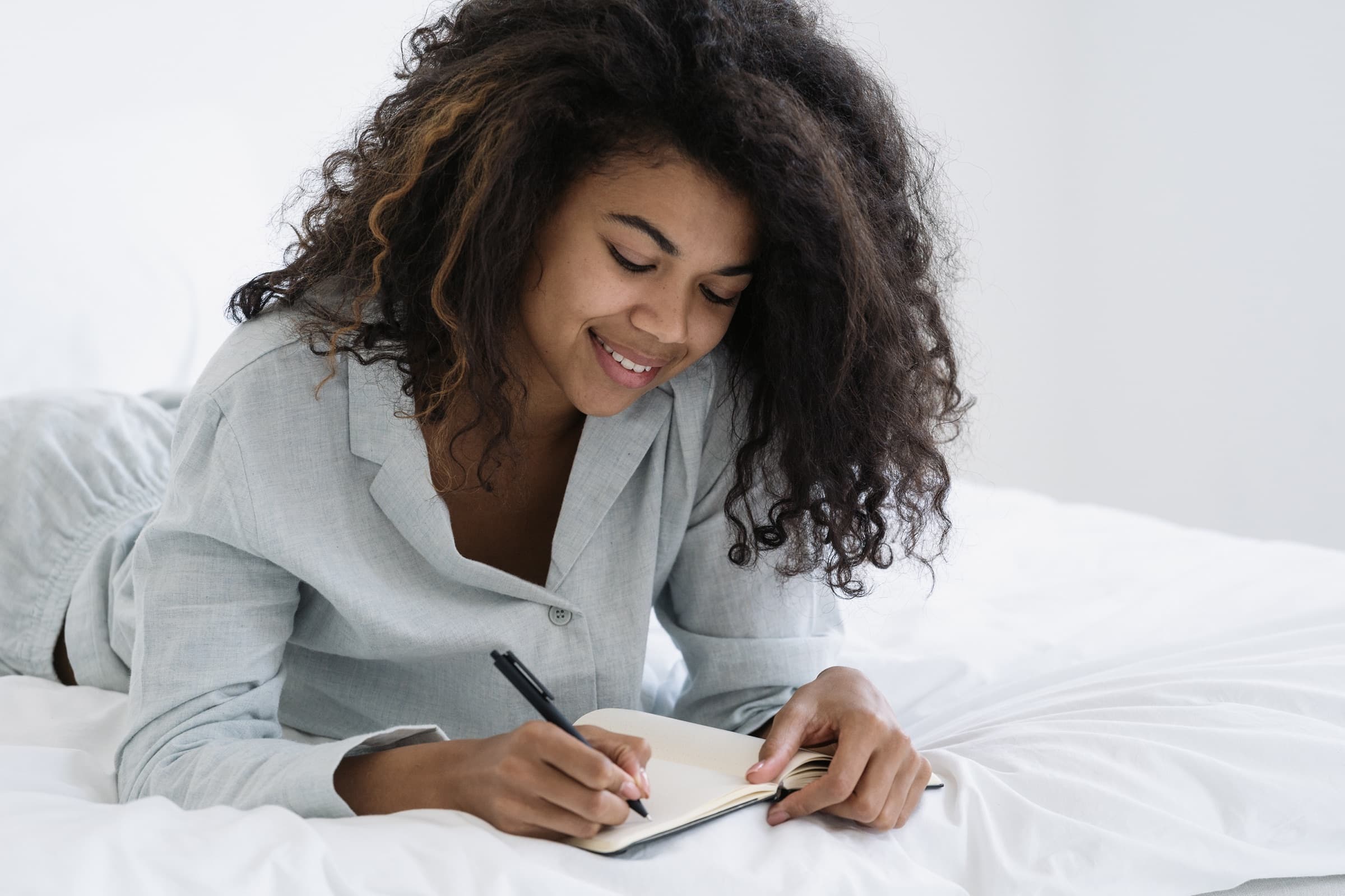A woman in pyjamas lying on her bed, writing in a notebook with a pen.